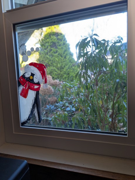 A cartoon penguin with his eyes closed is on a window pane. A garden can be seen through the window